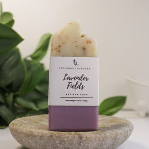 Bar of soap labeled 'Lavender Fields' by Leelanau Lavender on a stone surface with green leaves in the background.